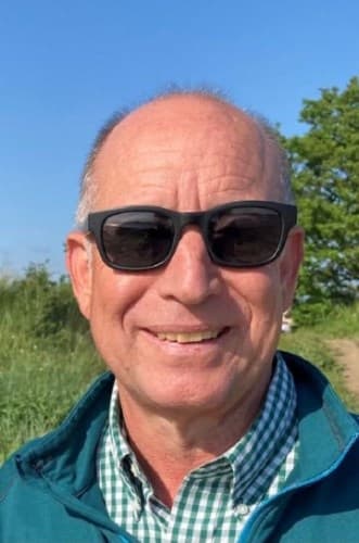 Joe wears sunglasses and smiles. He is outside, and trees and greenery are in the background.