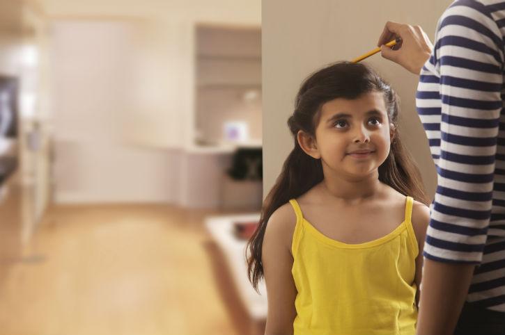 A young brown-skinned girl smiles up at someone using a pencil to measure her growth on a wall.