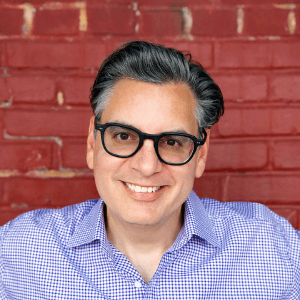 A close-up of Thomas standing in front of a brick wall. He has salt-and-pepper hair, wears glasses, and wears a purple gingham dress shirt.