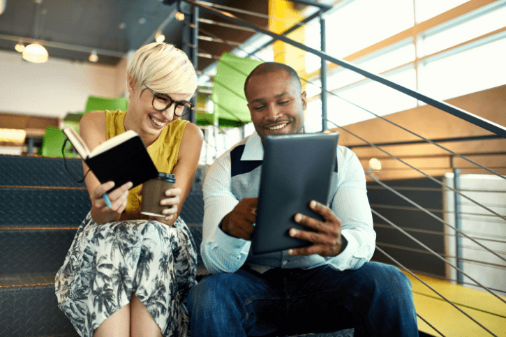 A man shows a tablet to a woman holding coffee and a notebook