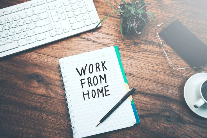 An open spiral-bound pad with, "WORK FROM HOME" on the page. Around it is a keyboard, a plant, glasses, and a cup of coffee.