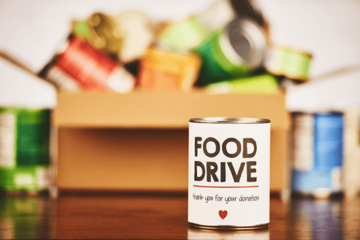 A can labeled "Food Drive" with a red heart. Behind it is a blurred box of canned goods.