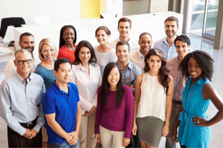 Fifteen people dressed in business casual stand in an office lobby and look up, smiling towards the camera.