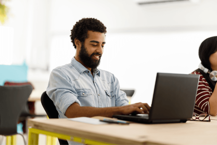 A bearded man works on a laptop while sitting at a wooden conference room table.