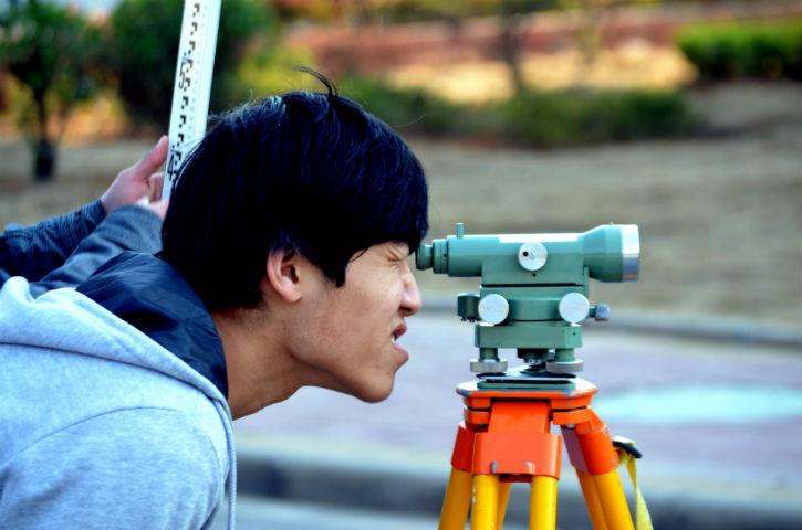 A man of Asian descent peers through a surveying tool