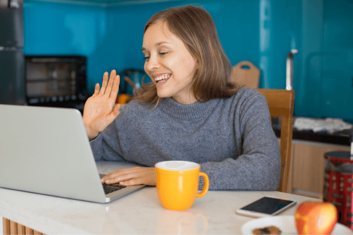 A woman sits in her kitchen and waves to her laptop camera