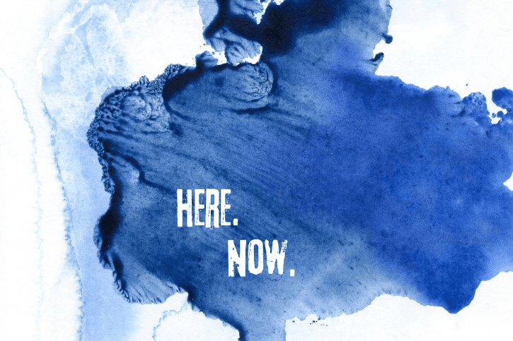 The words "Here" and Now" in white on a splotch of blue set against a white background.