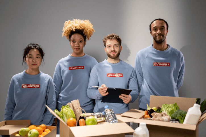 Four people stand next to one another looking into the camera. They're wearing grey sweatshirts with the word VOLUNTEER in white type on a red background. In front of them is a table filled with boxes of vegetables and water bottles.