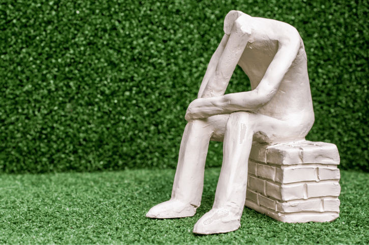 A toy-sized statue of headless person sitting on a brick wall. It sits in front of a fake green hedge.