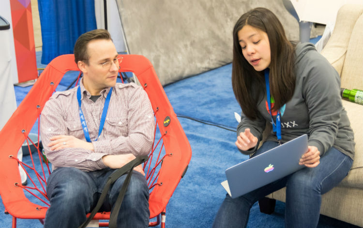 2 people, seated and talking; one person is showing the other something on a laptop
