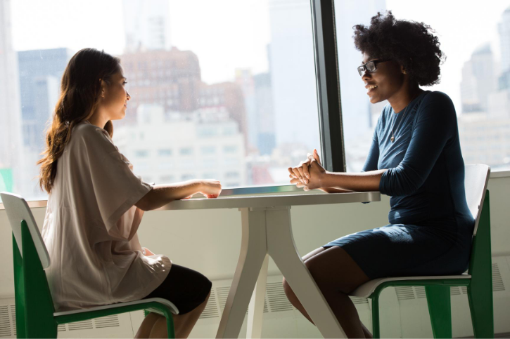 Two women sit across from each other at a small round table against a large office window.