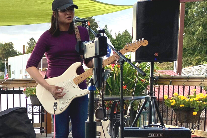 Samara Islam wears a black baseball cap and a purple long sleeve shirt, holds a white guitar and sings into a microphone.